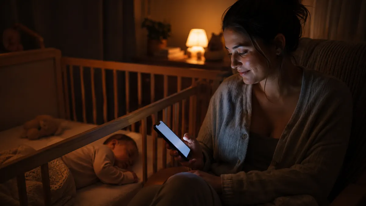 A mother checking her phone beside her sleeping baby at night.
